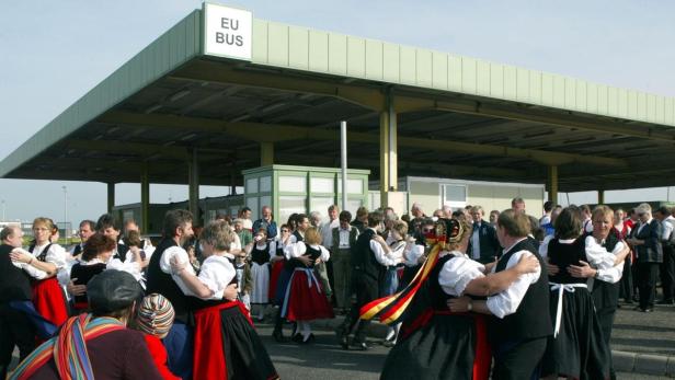 Menschen in Tracht tanzen vor einem ehemaligen Grenzübergang mit der Aufschrift „EU BUS“.