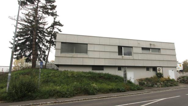 Ein modernes, zweistöckiges Gebäude mit grauer Fassade und rechteckigen Fenstern steht an einer Straße.