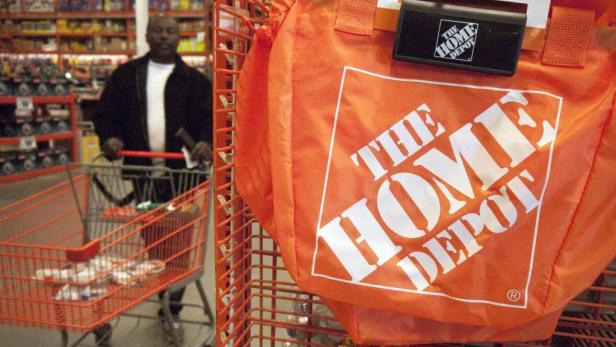 Eine orangefarbene Einkaufstasche von The Home Depot hängt an einem Regal, während ein Kunde im Hintergrund einkauft.