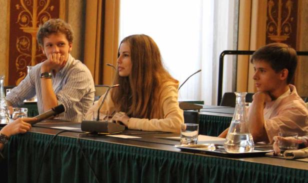 Drei junge Leute sitzen an einem Tisch mit Mikrofonen, möglicherweise bei einer Konferenz.