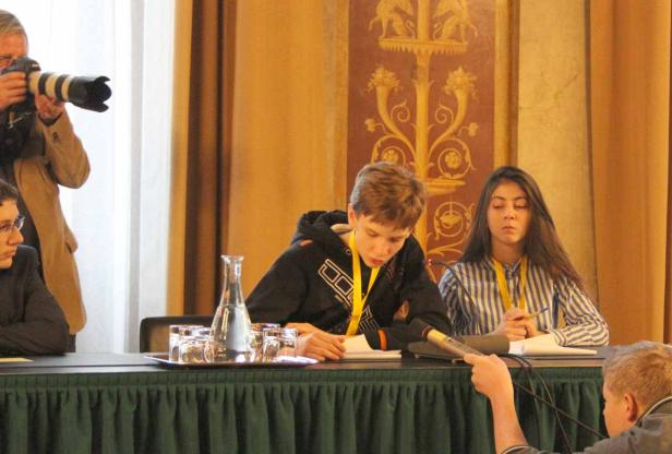 Zwei Jugendliche sitzen an einem Tisch mit Mikrofonen, während ein Fotograf im Hintergrund steht.