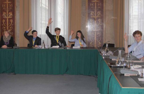 Fünf Jugendliche sitzen an einem Tisch und heben ihre Hände.