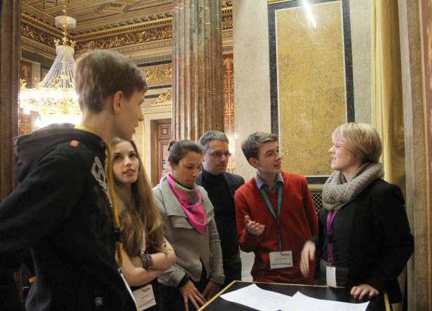 Eine Gruppe von Personen steht in einem prunkvollen Raum und diskutiert.