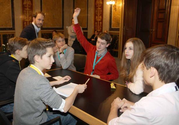 Eine Gruppe Jugendlicher sitzt an einem Tisch, ein Junge hebt die Hand.