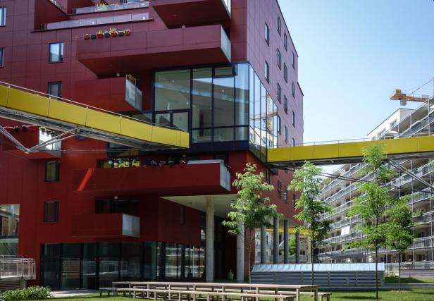 Ein modernes, rotes Gebäude mit gelben Übergängen und begrünten Balkonen.