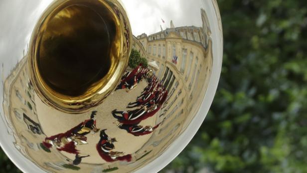 In der Tuba spiegelt sich eine Militärkapelle vor einem Gebäude mit französischer Flagge.