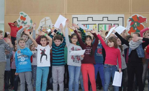 Eine Gruppe von Schulkindern steht auf einer Bühne und singt mit erhobenen Händen.