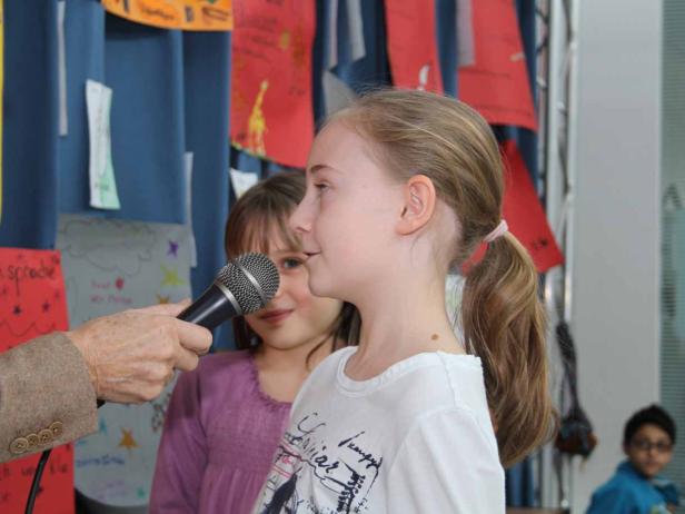 Ein Mädchen spricht in ein Mikrofon, während ein anderes Mädchen im Hintergrund lächelt.