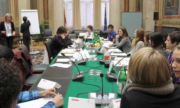 Eine Gruppe junger Leute sitzt an einem langen Tisch mit Mikrofonen in einem Konferenzraum.