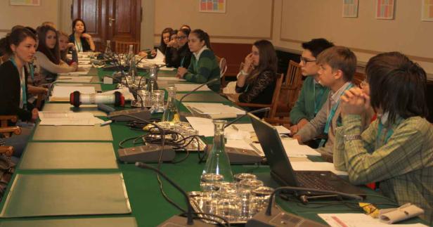 Eine Gruppe junger Leute sitzt an einem langen Tisch mit Mikrofonen und Laptops.