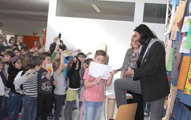 Ein Musiker spielt Gitarre vor einer Gruppe von Kindern, von denen einige Zeichnungen hochhalten.
