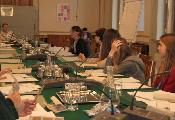 Mehrere Personen sitzen an einem langen Tisch mit Mikrofonen bei einer Konferenz.