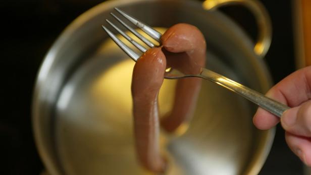 Eine Person hält eine Wurst mit einer Gabel über einen Topf mit Wasser.