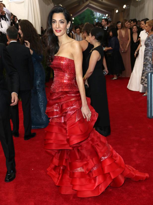 Amal Clooney in einem roten, gestuften Kleid auf einem roten Teppich.