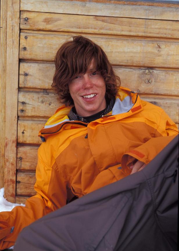 Ein junger Mann mit roten Haaren und Sommersprossen lächelt in einer orangefarbenen Jacke vor einer Holzwand.