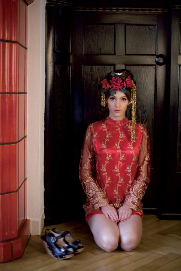 Eine Frau kniet in einem roten Kleid mit Blumenkopfschmuck vor einer dunklen Tür.