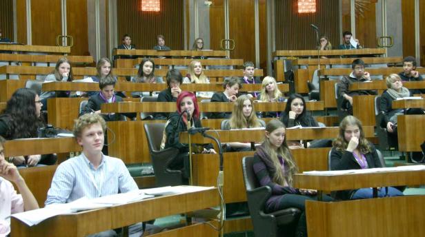 Jugendliche sitzen in einem großen Saal mit hölzernen Bänken und Tischen.