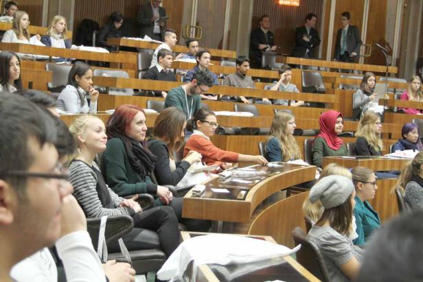 Zahlreiche Menschen sitzen in einem großen Hörsaal mit hölzernen Bänken.