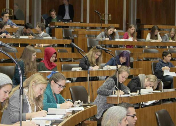 Schüler sitzen in einem Klassenzimmer mit Pulten und Mikrofonen und schreiben.