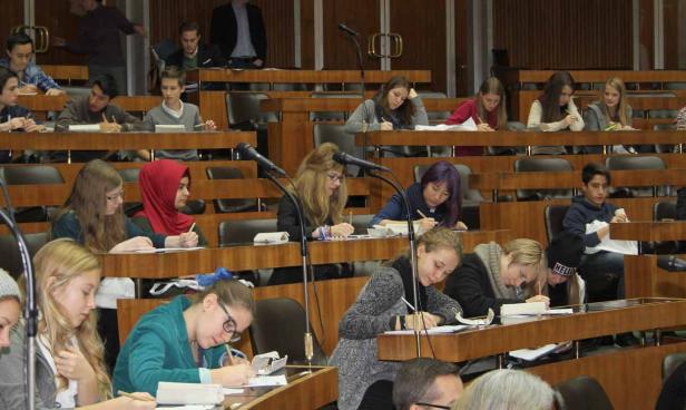 Schüler sitzen in einem Klassenzimmer mit hölzernen Tischen und schreiben.