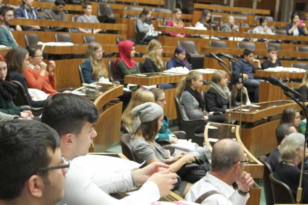 Zahlreiche Menschen sitzen in einem Hörsaal mit hölzernen Tischen und Stühlen.