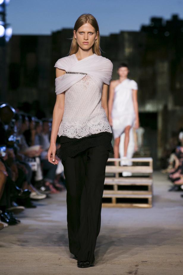 Eine Frau präsentiert ein weißes Oberteil mit Spitze und eine schwarze Hose auf dem Laufsteg.