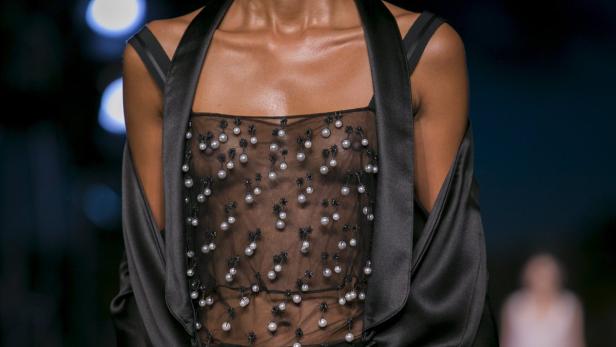 Eine Frau trägt ein transparentes Oberteil mit Perlenapplikationen und einen schwarzen Satinmantel.