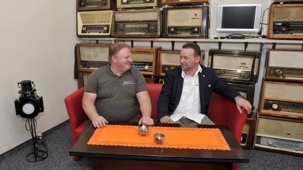 Zwei Männer sitzen auf einem roten Sofa vor einer Wand mit alten Radios.