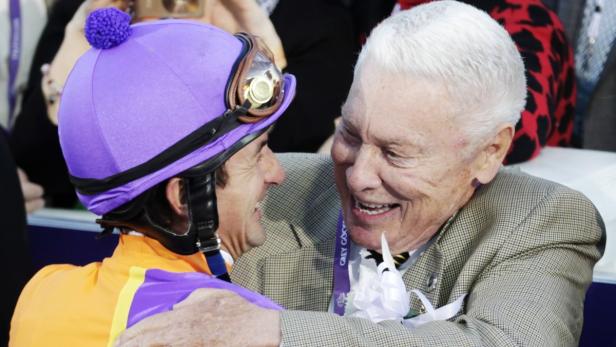 Ein Jockey in lila und orangefarbener Rennkleidung umarmt einen älteren Mann in einem karierten Sakko.