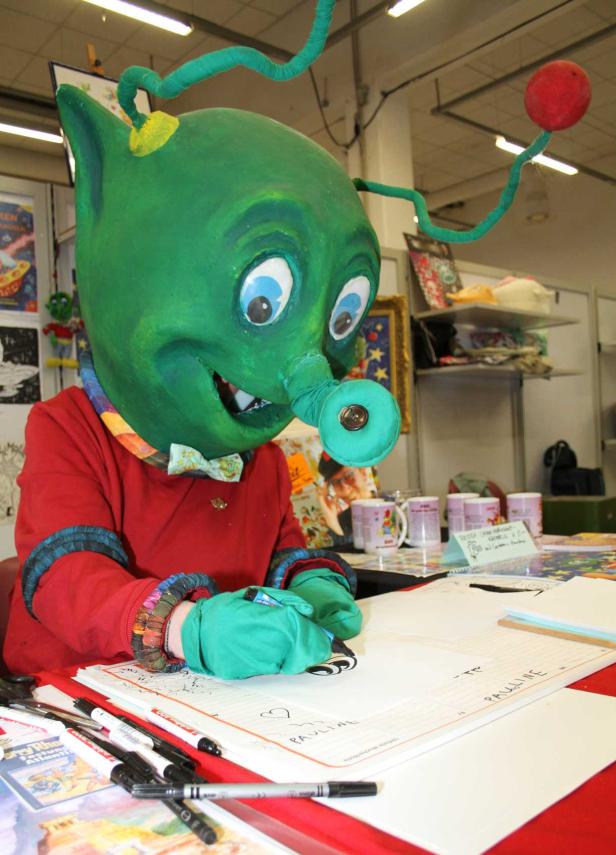 Eine Person in einem grünen Alienkostüm zeichnet auf einem Blatt Papier.