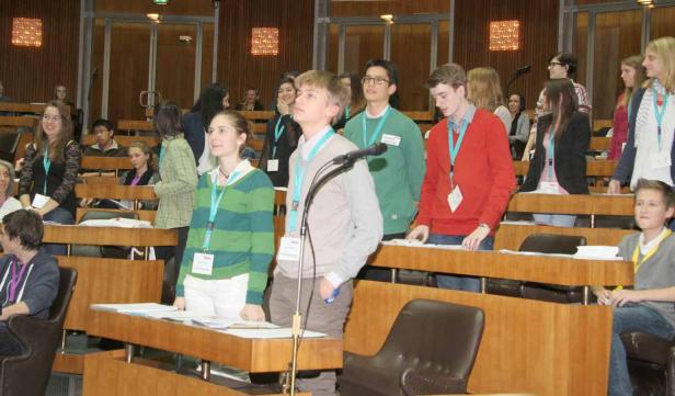 Jugendliche stehen und sitzen in einem holzvertäfelten Raum mit Tischen und Mikrofonen.