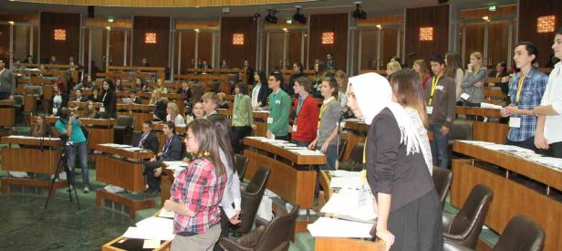 Eine Gruppe junger Menschen sitzt oder steht in einem großen Saal mit Holzbänken.