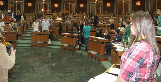 Eine Gruppe junger Menschen sitzt in einem großen Saal mit hölzernen Tischen und grünen Teppichen.