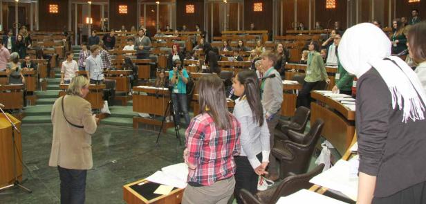Menschen sitzen und stehen in einem großen Saal mit Holzbänken.