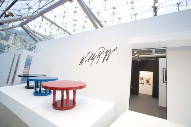 Ein Ausstellungsraum mit farbigen Tischen und dem Schriftzug „Willy Rizzo“ an der Wand.