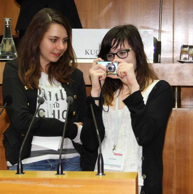 Zwei junge Frauen stehen an einem Rednerpult, eine fotografiert mit einer kleinen Kamera.