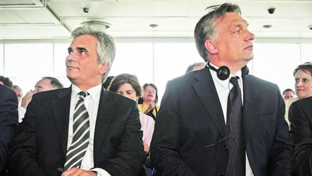 Zwei Männer in Anzügen sitzen in einer Menschenmenge und schauen in die Ferne.