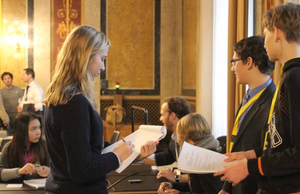 Eine junge Frau macht sich Notizen, während andere Personen im Hintergrund stehen oder sitzen.
