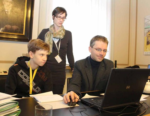 Ein Mann arbeitet am Laptop, während ein Jugendlicher und eine Frau zusehen.