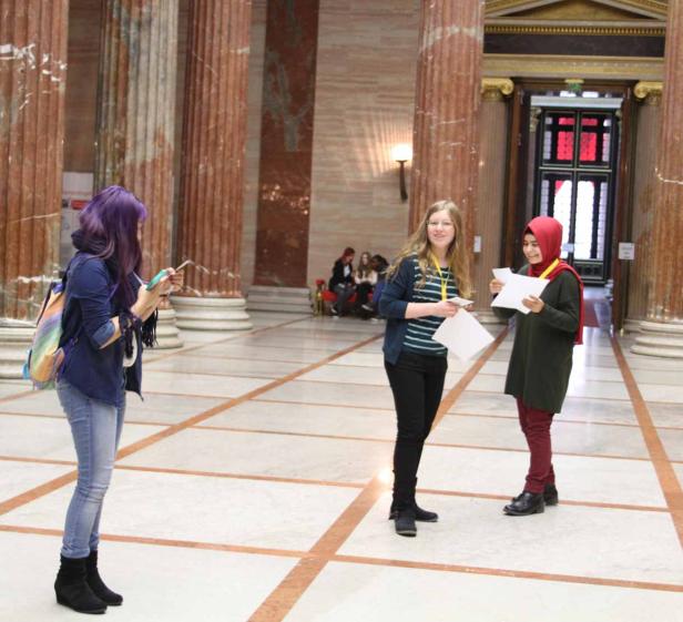 Drei junge Frauen stehen in einer Halle mit Säulen, eine fotografiert mit dem Handy.