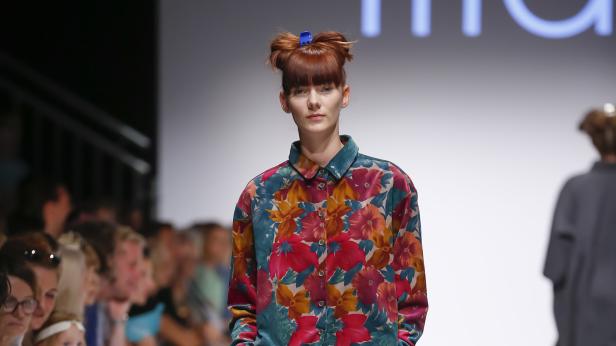 Eine Frau mit hochgesteckten roten Haaren und blauer Haarspange trägt ein buntes Blumenhemd.