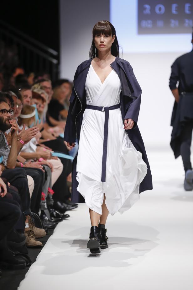 Eine Frau in einem weißen Kleid und einer dunklen Jacke läuft auf dem Laufsteg.