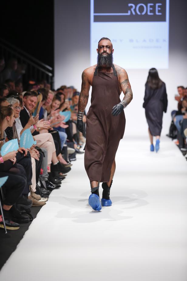 Ein tätowierter Mann mit Bart in einem braunen Overall auf einem Laufsteg.