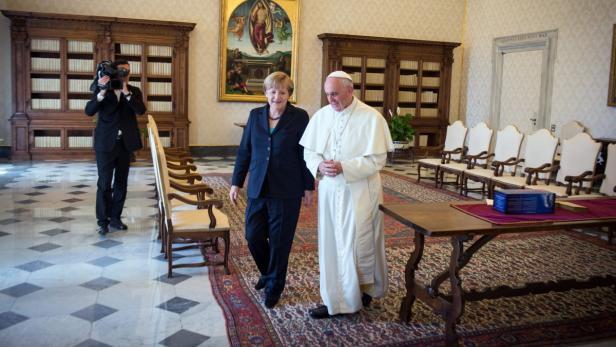 Angela Merkel und Papst Franziskus gehen in einem formellen Raum nebeneinander her.