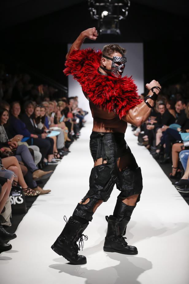 Ein muskulöser Mann mit einer silbernen Maske und einem roten Federkragen auf dem Laufsteg.
