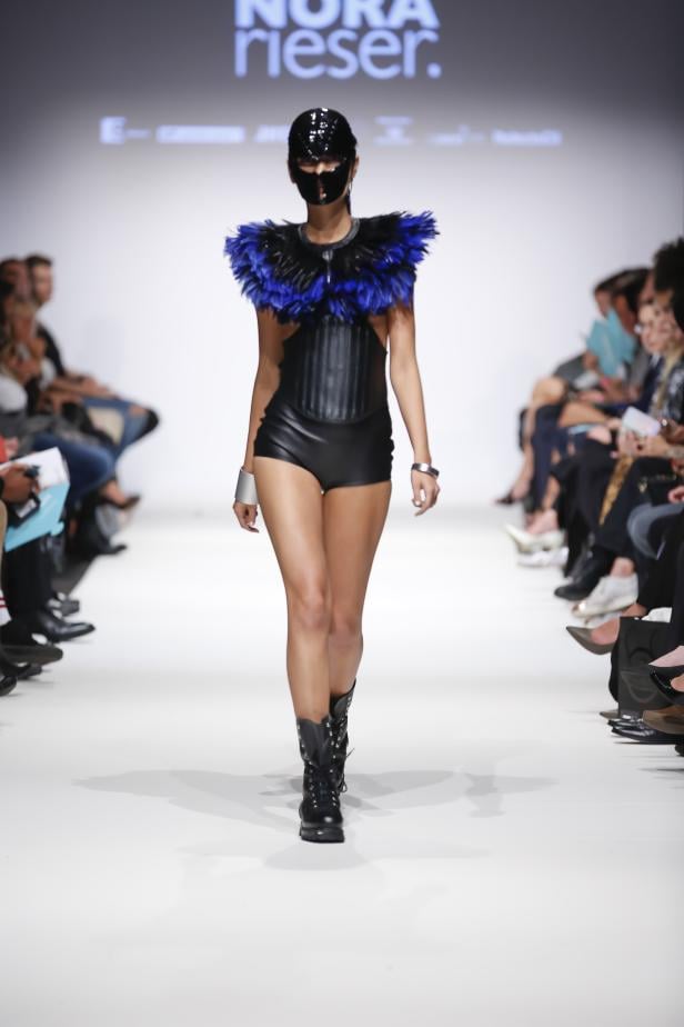 Eine maskierte Frau in einem schwarzen Lederbody mit blauen Federn auf einem Laufsteg.