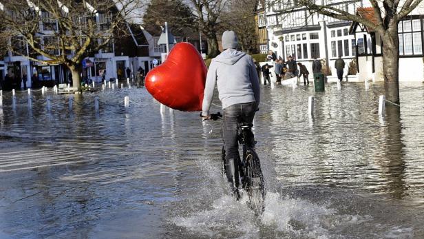 Ein Mann fährt mit dem Fahrrad durch eine überflutete Straße und hält einen roten Herzluftballon.