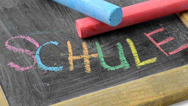 Die Tafel zeigt das Wort „Schule“, geschrieben mit bunter Kreide.