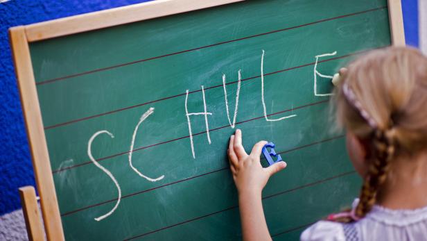 Ein Kind schreibt mit Kreide „Schule“ auf eine grüne Tafel.