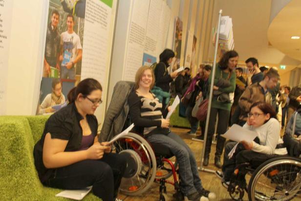 Mehrere Personen betrachten Dokumente auf einer Veranstaltung, zwei davon im Rollstuhl.